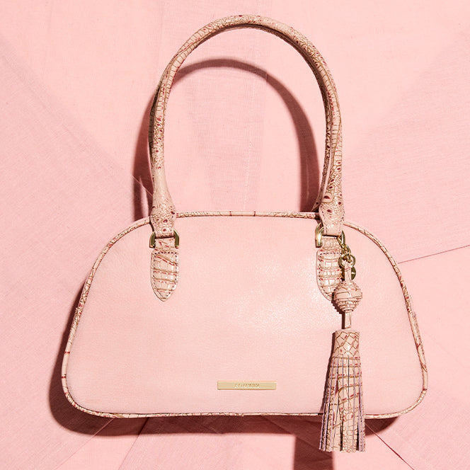 Light pink leather satchel handbag with rounded top handles, crocodile embossed trim, gold hardware, and a matching tassel charm, displayed against a soft pink background.