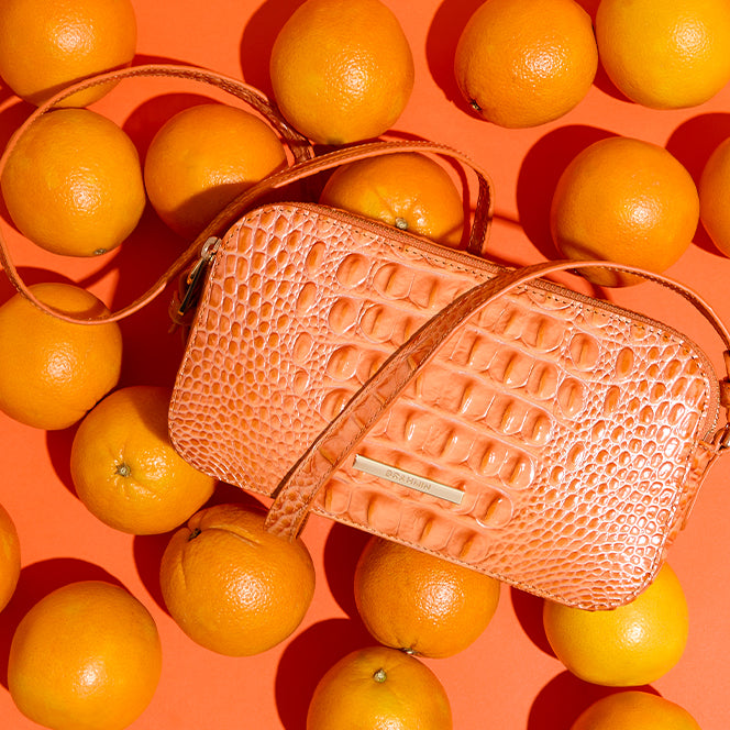 Orange croc-embossed leather crossbody bag with zip-top closure and adjustable strap, styled among fresh oranges