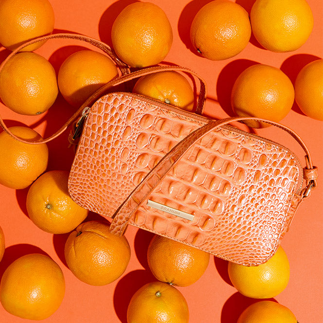 Coral orange crocodile embossed crossbody bag displayed on an orange background surrounded by whole oranges