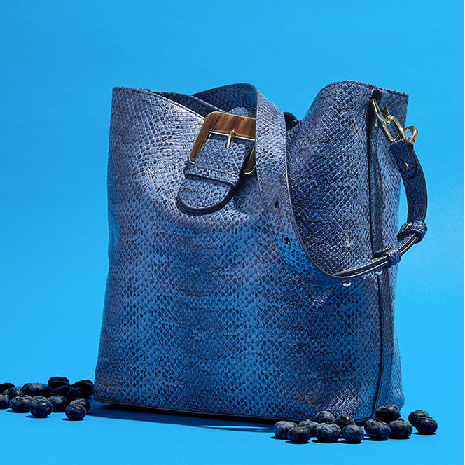 Blue textured leather bucket bag with adjustable shoulder strap and wide front buckle detail on a blue backdrop
