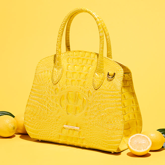 Bright yellow crocodile embossed satchel displayed on a yellow background with whole and sliced lemons