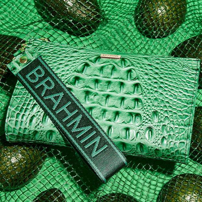 Emerald green crocodile embossed wristlet with a black Brahmin logo strap displayed on green netting with avocados