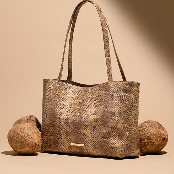 A tall shopper tote bag crafted from natural brown snakeskin textured leather with long shoulder straps. The bag sits on a beige surface flanked by three whole coconuts.