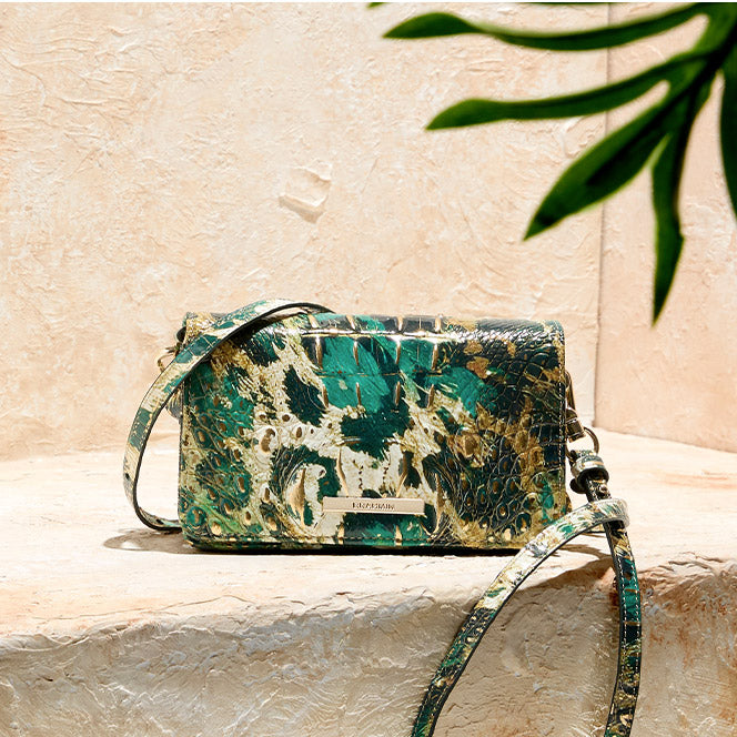 Teal, cream and gold Brahmin crossbody bag on a stone ledge with palm leaves overhead