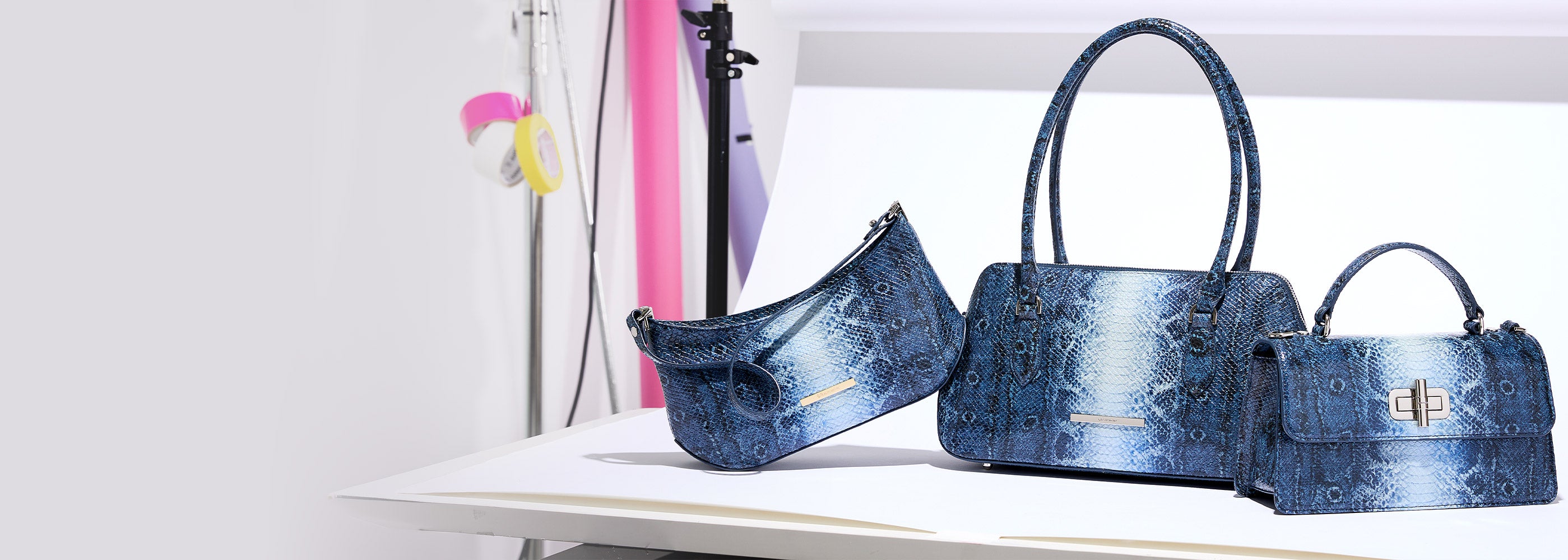 Three metallic blue snake embossed Brahmin handbags displayed on white studio blocks, featuring a large double handle satchel, a curved zip top shoulder bag, and a small structured top handle bag with a silver turn lock.