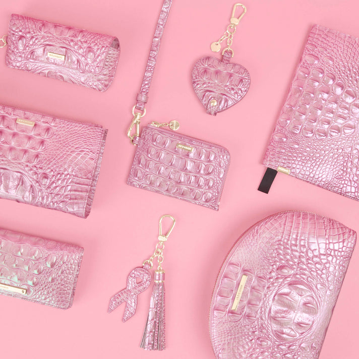 Collection of glossy pink croc embossed accessories arranged on a pink background, including wallets, zip pouches, a wristlet, heart shaped charm, and ribbon and tassel keychain, all finished with gold Brahmin hardware and logo details.