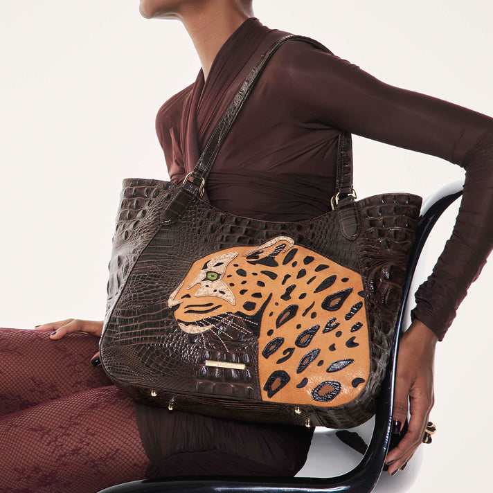 Model carrying a dark brown Brahmin designer tote bag with signature embossed texture, double shoulder straps, gold tone hardware and a large tan leopard graphic on the front.
