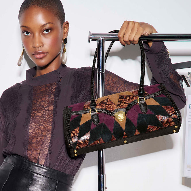 Model holding a structured Brahmin satchel featuring geometric patchwork in burgundy, tan, forest green, and snakeskin inspired textures with black croc embossed handles and gold hardware.