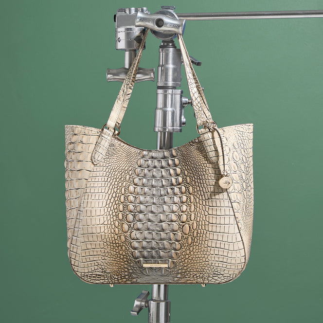 Large cream and taupe Brahmin tote in croc embossed leather with two shoulder straps, a spacious curved silhouette, gold logo hardware, and dimensional crocodile texture against a green background.