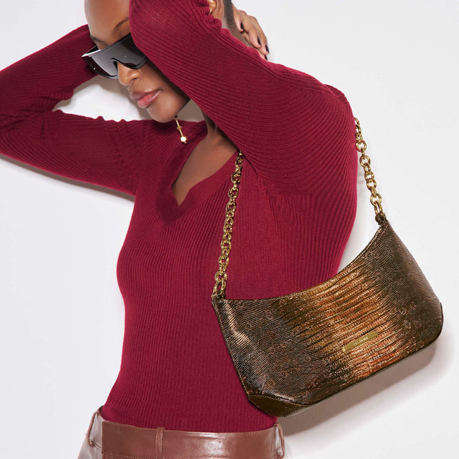 Model in a deep red ribbed top carrying a metallic bronze Brahmin shoulder bag with a curved silhouette, textured leather finish, gold chain strap, and gold logo hardware.