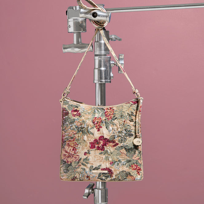 Brahmin shoulder bag featuring a cream botanical floral print with burgundy flowers and green foliage, croc embossed texture, slim shoulder strap, and gold logo hardware against a dusty pink background.