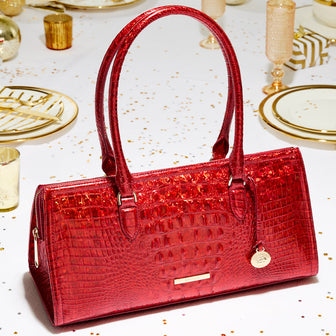 Close-up of a red Brahmin satchel Sophia handbag in crocodile-embossed leather with glossy finish, rectangular structured body, double top handles, zip closure, and signature round gold medallion charm, styled on a holiday table with gold-rimmed plates, candles, and festive confetti