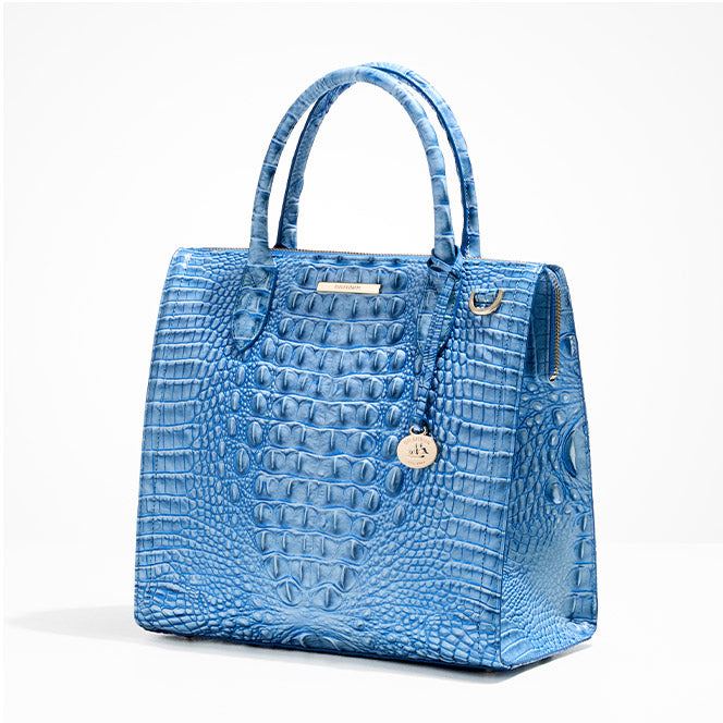 Bright blue croc-embossed leather satchel with a structured boxy shape, zip-top closure, double rolled handles, gold hardware, and a round gold logo charm on a white background.