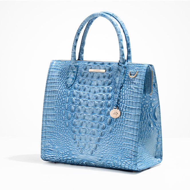 Structured light-blue crocodile-embossed leather satchel with rounded top handles, zip-top closure, and a hanging round logo charm on a white background.