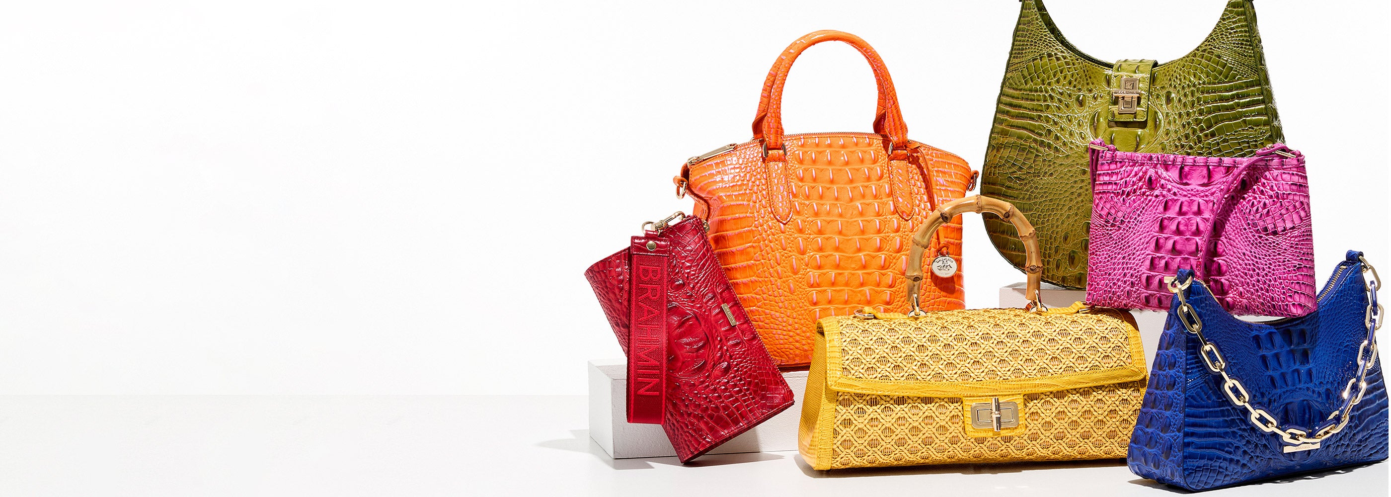 Colorful assortment of Brahmin handbags in bold croc embossed leather styles, featuring a red wristlet, orange satchel, yellow woven flap clutch with bamboo handle, olive green shoulder bag, pink drawstring crossbody, and royal blue shoulder bag with chunky chain strap on a white studio background.