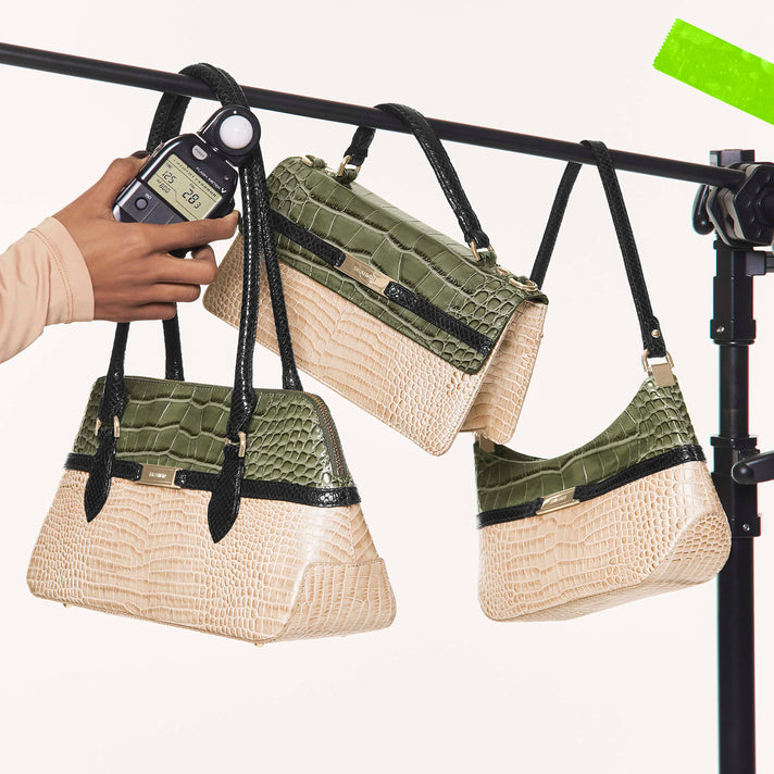 Three Brahmin handbags in cream, sage green, and black croc embossed leather, including a structured tote, flap shoulder bag, and curved shoulder bag, displayed hanging from a studio rack.