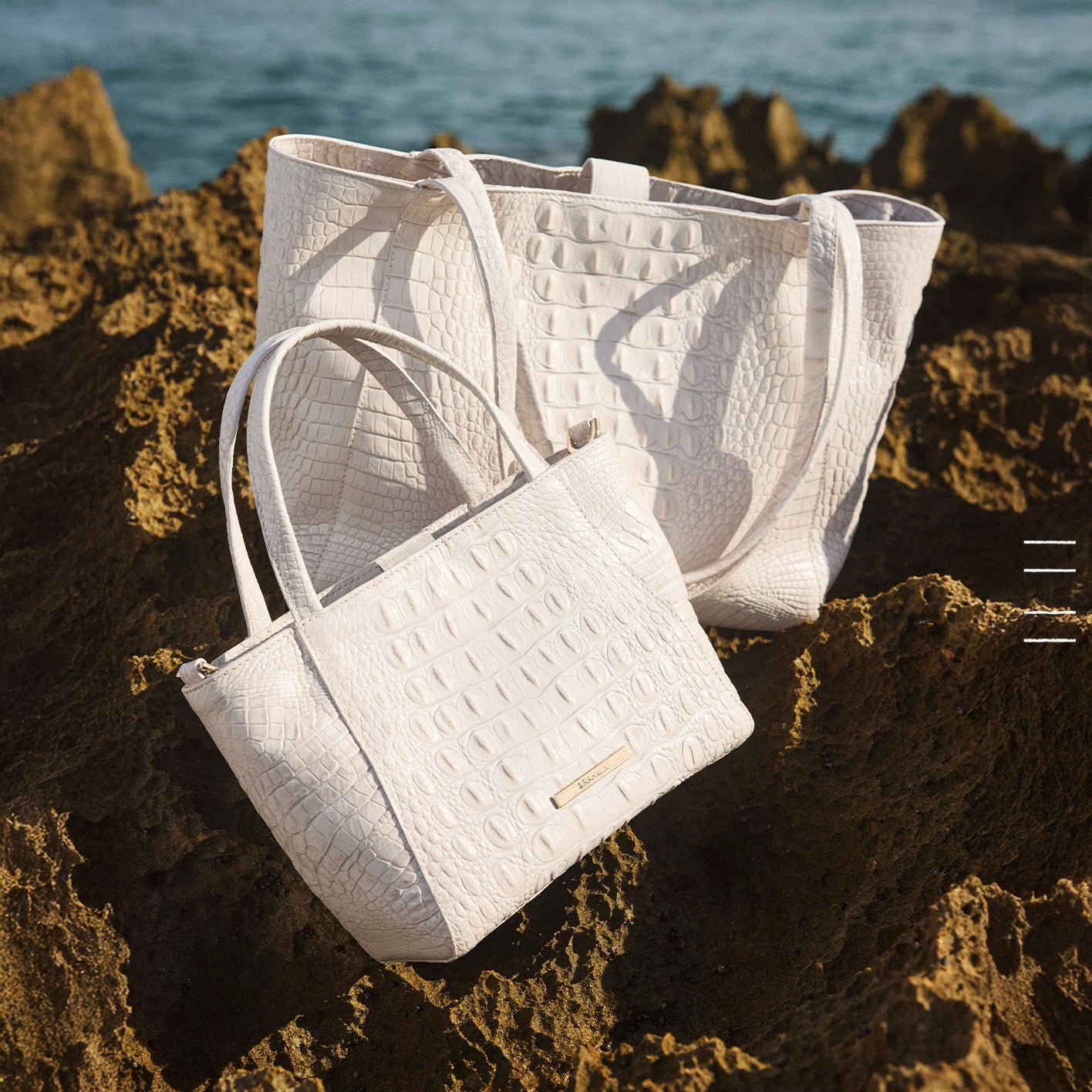 Two white croc-embossed leather tote bags with structured silhouettes and long shoulder straps displayed on rocky shoreline near the ocean