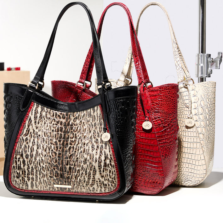 Three Brahmin shoulder totes arranged in a studio display, featuring a black framed bag with an ivory animal inspired center panel, a bright red tote, and a cream tote, all finished in embossed leather with round logo charms.