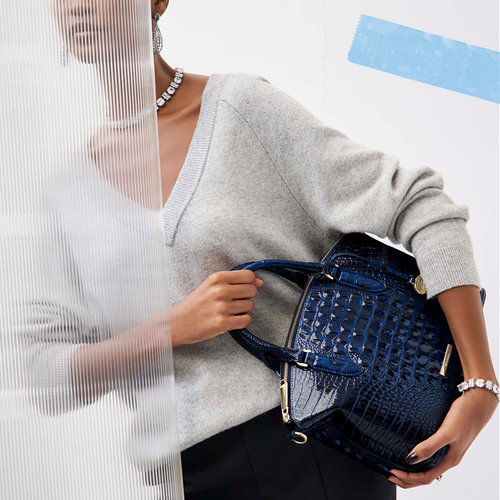 Deep navy blue croc embossed leather satchel with a structured rounded silhouette, pronounced glossy crocodile scales, two rolled top handles, gold zipper and hardware, and a slim gold Brahmin logo bar, held against a model wearing a light gray sweater.