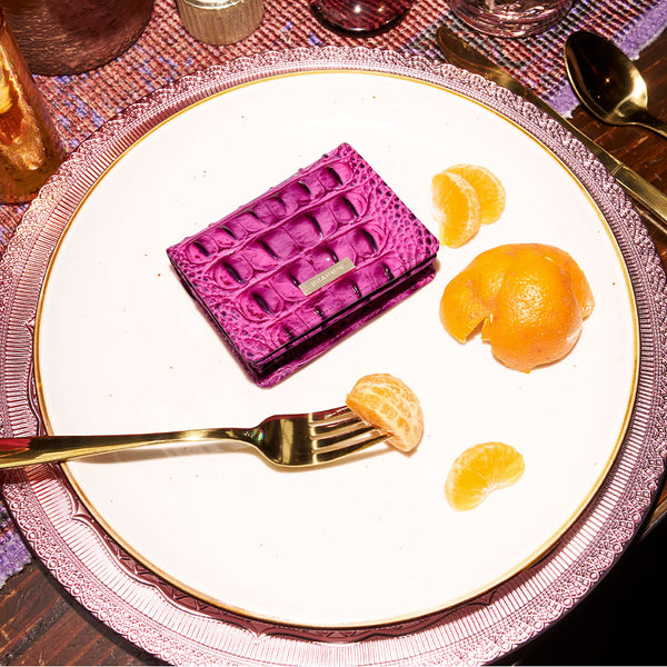 Pink Brahmin croc embossed leather flap wallet with gold logo plaque, styled on a white plate with citrus accents