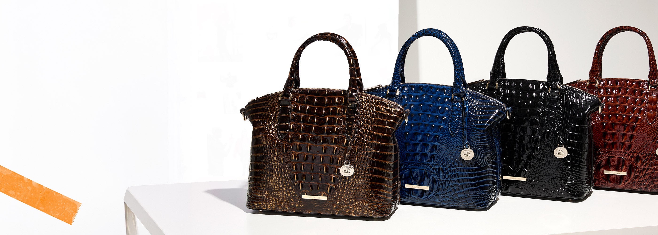 Four structured Brahmin croc embossed handbags arranged in a row in rich brown, deep blue, black, and reddish brown, each with rounded handles, gold logo plates, and Brahmin medallion charms.