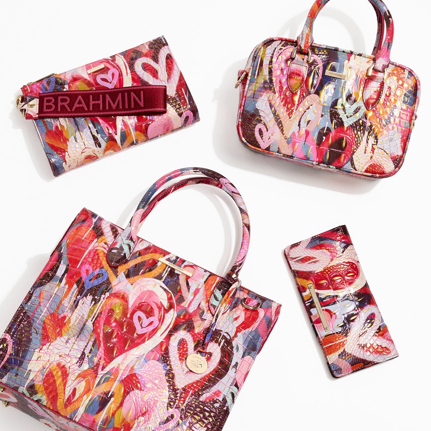 Flat lay of four accessories in a bold multicolor croc-embossed print: a large tote with round handles and a hanging charm, a small top-handle satchel, a long wallet, and a zip pouch with a wide ‘BRAHMIN’ strap, all on a white background.