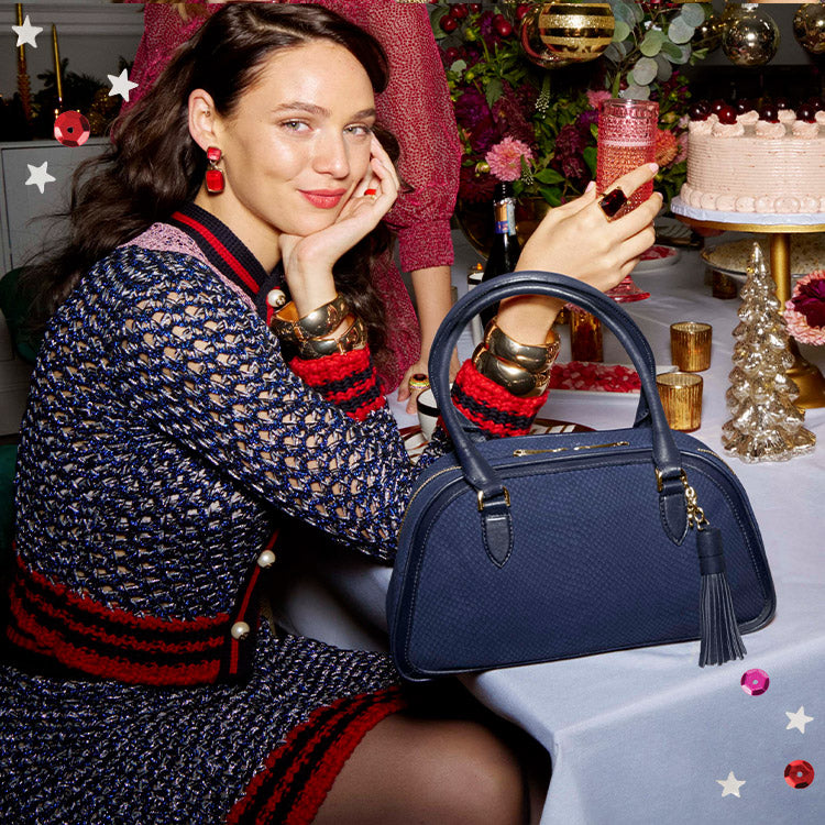 Woman in a navy knit dress with red trim sits at a festive holiday dinner table, resting her chin on her hand while a navy blue Brahmin satchel in embossed leather with dome silhouette, double rolled handles, top zip closure, and oversized tassel accent sits prominently in front of her on the table