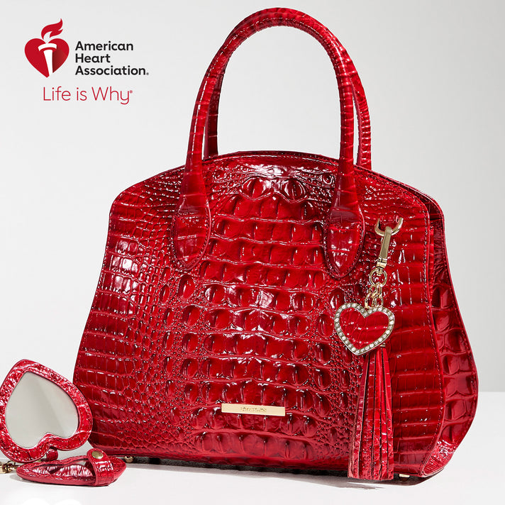 High-shine red crocodile-embossed leather satchel handbag with structured silhouette, rolled top handles, gold logo bar, and detachable heart-shaped charm tassel. Shown with American Heart Association Life Is Why logo