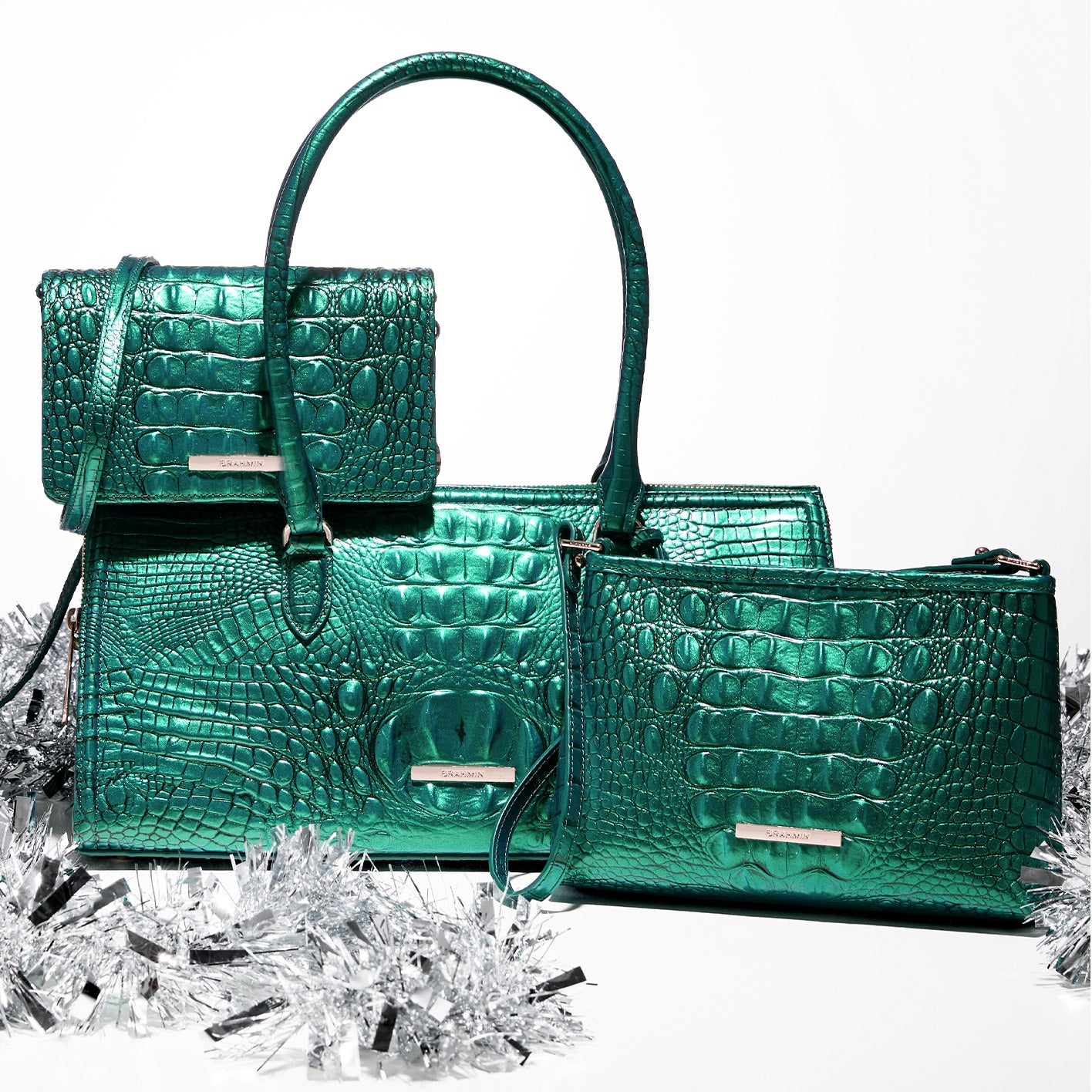 Trio of metallic green Brahmin handbags—Sophia satchel, Minuette shoulder bag, and Lorelei crossbody—styled together with silver holiday tinsel.