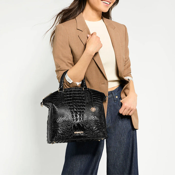 Model wearing a tan blazer and dark denim while carrying a glossy black Brahmin crocodile embossed satchel.