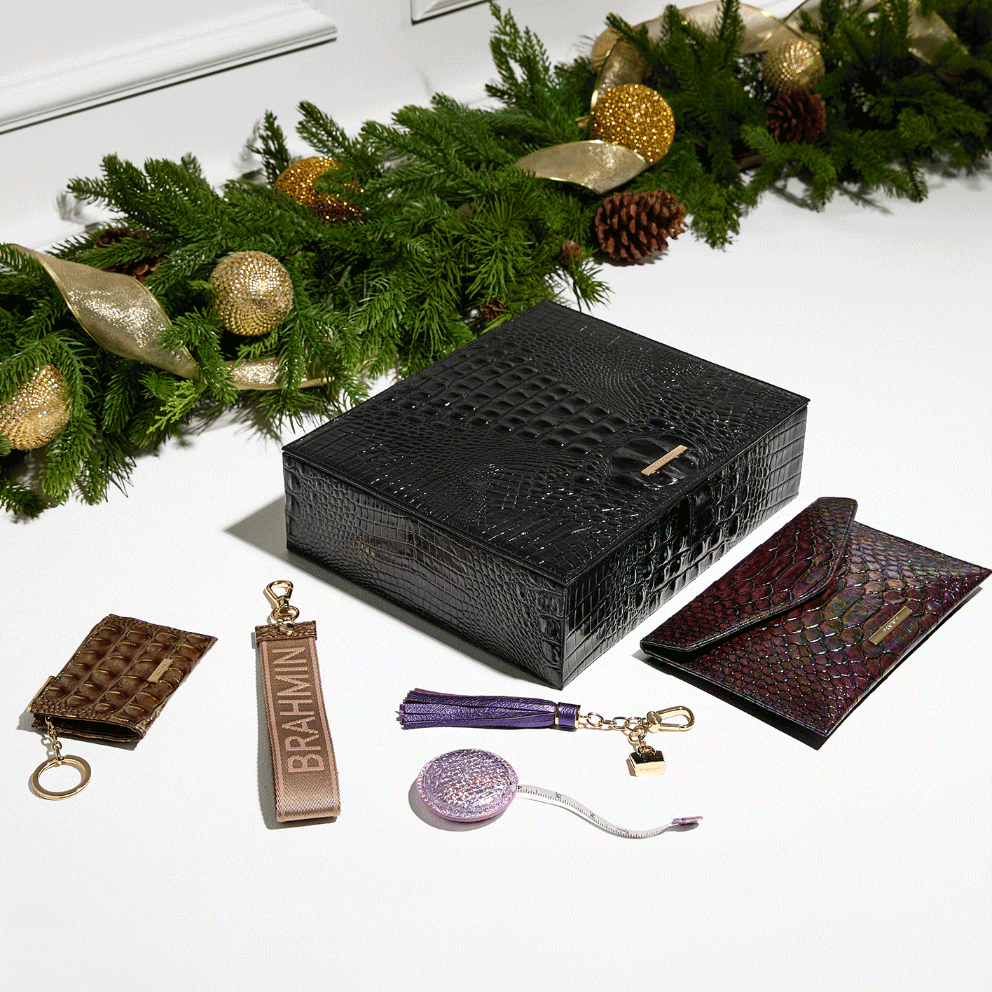 Animated holiday GIF showcasing Brahmin’s luxury leather gift set. A black embossed Melbourne jewelry box is surrounded by coordinating leather accessories, including an iridescent envelope clutch, brown croc keychain, pink logo web strap, purple tassel charm, and a lavender tape measure key fob. Displayed beside festive greenery with gold ornaments and pinecones, this elegant $515 collection highlights Brahmin’s signature craftsmanship and makes the perfect holiday gift.