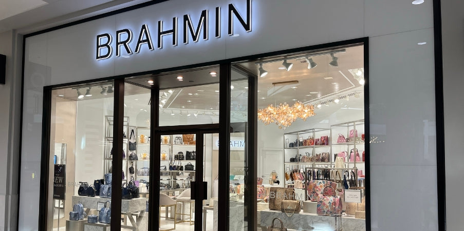 Brahmin handbag displayed in a modern retail setting, featuring a structured silhouette, embossed texture, gold-tone hardware, top handle and shoulder strap, shown on a glass shelf with accessories in the background.