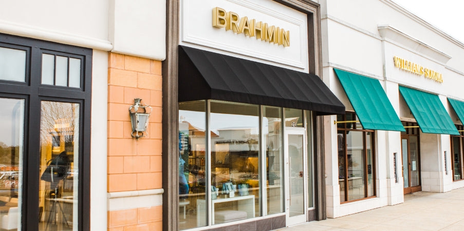 Brahmin handbag displayed with a structured silhouette, top handles, gold-tone hardware, and signature textured pattern; the bag features neutral tan accents and is shown upright on a shopping plaza sidewalk.