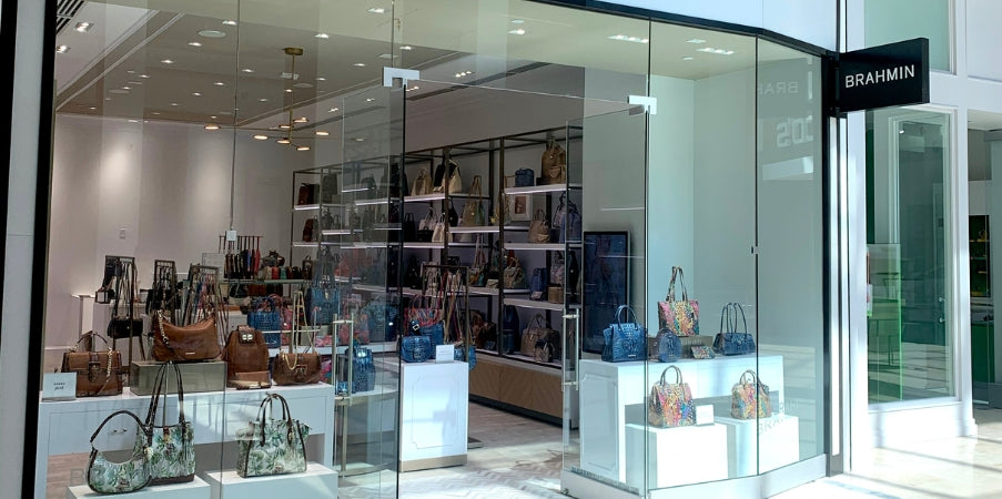 Brahmin handbags displayed on glass shelves and stands in a bright, modern store. Various purse silhouettes feature assorted colors, patterns, textured finishes, gold hardware, top handles, and crossbody straps.