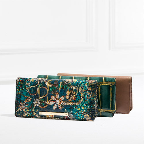 Trio of Brahmin leather wallets in embellished teal, green croc, and smooth taupe, displayed in a layered holiday-inspired setting.