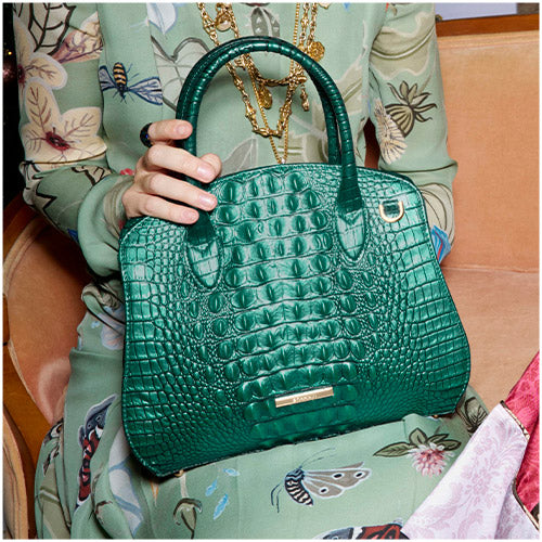 Woman holding a green Brahmin croc-embossed satchel, styled with a patterned holiday dress and gold jewelry.