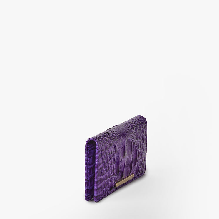 Brahmin purple embossed leather wallet with raised textured pattern, metallic logo plate on front, compact square silhouette standing upright on white background, no strap or visible interior details shown.