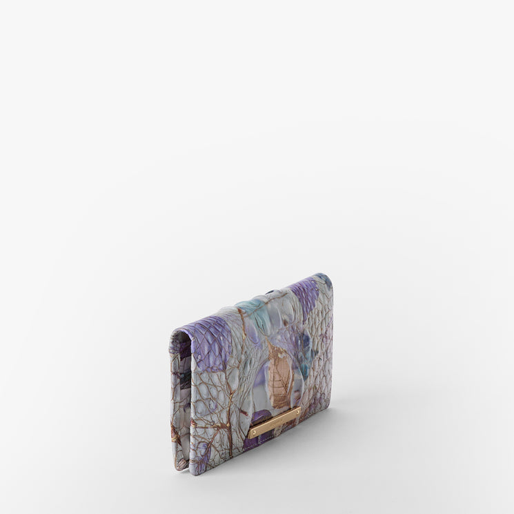 Brahmin folded wallet in pastel purple, blue, and beige with an abstract pattern, small gold-tone hardware accent at the bottom edge, shown on a white background. Silhouette and front details clearly visible.