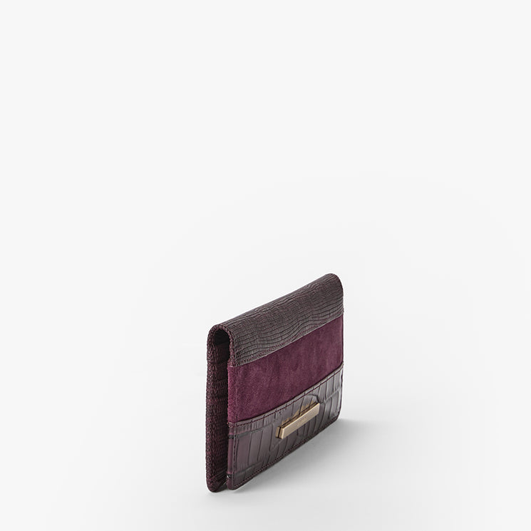 A slim bifold wallet featuring a dark plum textured crocodile-embossed leather exterior, a deep purple suede panel in the center, and a small gold-tone Brahmin plaque on the front, set against a plain white background.