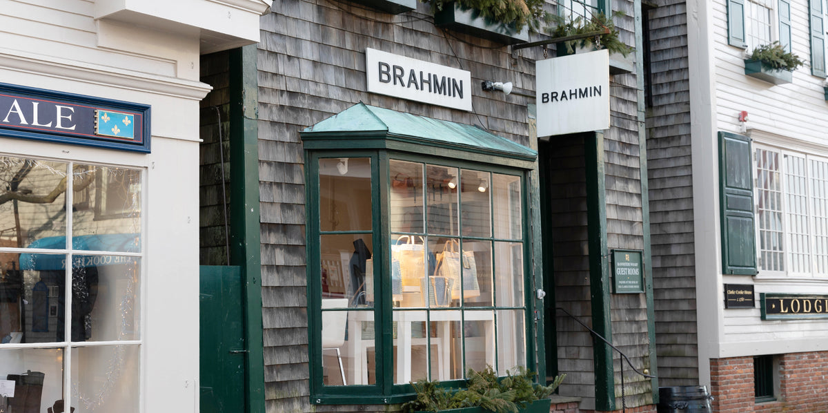 Brahmin storefront with green trim displays an assortment of handbags and totes in the front window beneath a Brahmin sign; neighboring shop signage and windows are visible on both sides.