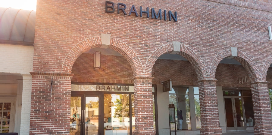 BRAHMIN handbag displayed in front of a red brick building, featuring bold branding. The bag showcases a structured silhouette, textured pattern, gold-tone hardware, top handles, and adjustable shoulder strap.
