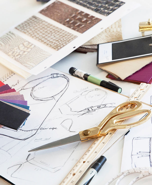 Brahmin handbag in a structured silhouette with embossed texture, gold hardware, top handles, and adjustable strap displayed on a desk with fabric swatches, rulers, sketches, and markers.
