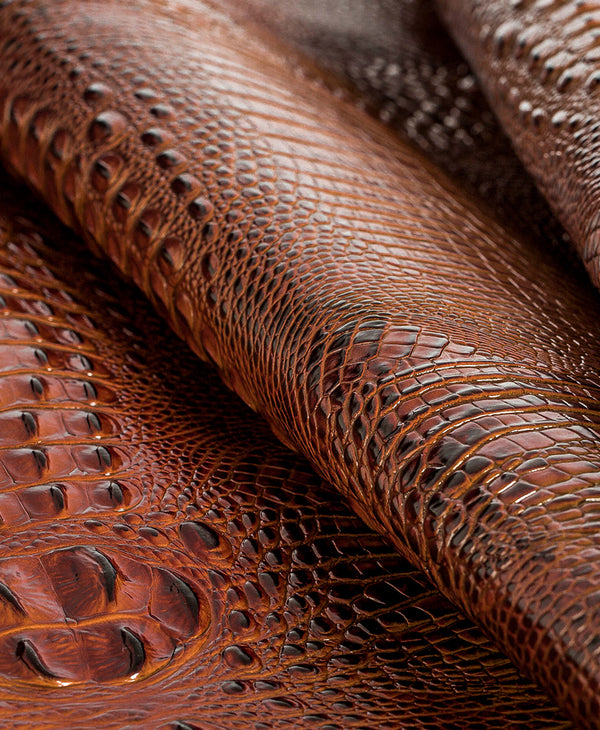 Brahmin handbag in rich brown with a textured, embossed pattern and glossy finish, featuring shiny gold hardware and visible natural folds. Close-up highlights signature scaled effect and premium craftsmanship.