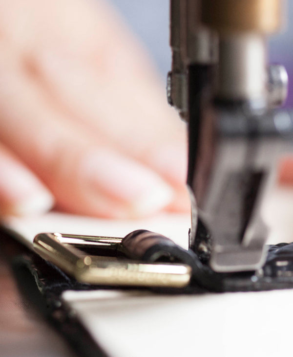 Brahmin crossbody bag in textured leather with gold-tone buckle hardware, shown in a close-up view as stitching is applied. The blurred hand in the background guides the material for precise detail work.