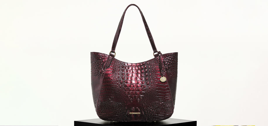 A burgundy crocodile-embossed leather tote bag with double shoulder straps and a small gold-tone hanging charm, displayed on a black pedestal against a white background.