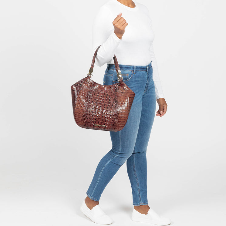 Model holds a large Brahmin handbag in textured brown finish with structured silhouette and top handles, styled on her arm. Bag features gold-tone hardware and signature Brahmin detailing, shown from a front view.