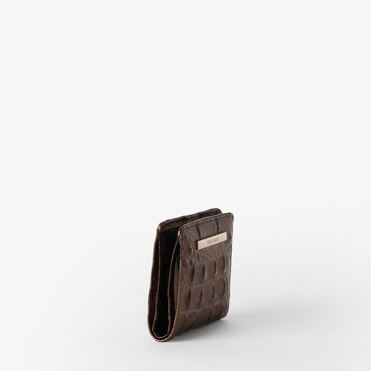 Brahmin brown embossed wallet with a distinctive textured pattern, upright on a white background. Features a metallic rectangular Brahmin logo plaque on the front; compact silhouette, no strap visible.