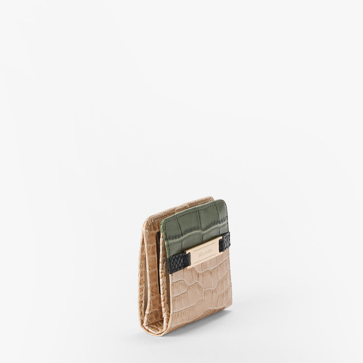 A small bi-fold wallet with a textured beige crocodile-patterned body, a dark olive green card pocket, and a black leather accent strap with a gold-tone Brahmin plaque, shown on a clean white background.