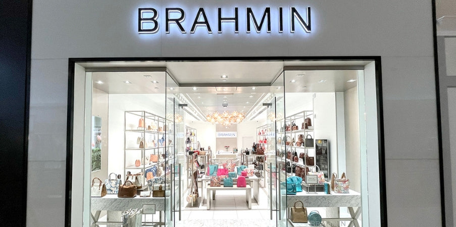 Brahmin handbag displayed on a shelf, showcasing a structured silhouette with top handles, gold-tone hardware, textured pattern, and adjustable strap; the bag is presented in front of a well-lit store interior.