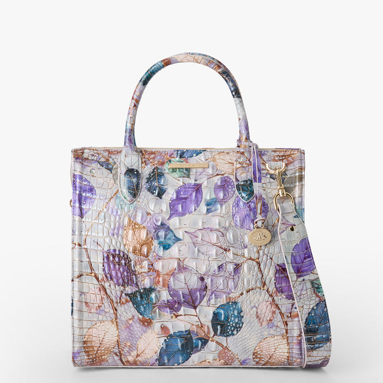 Brahmin structured satchel in a pastel floral and leaf print with purple, teal, and beige tones, textured finish, gold hardware, double top handles, detachable shoulder strap with gold charm; front view.